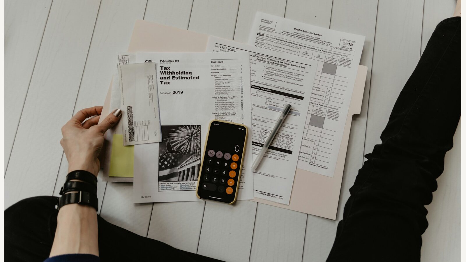 Tax forms spread on a white desk with a phone calculator open on top, a pen resting across the paperwork
